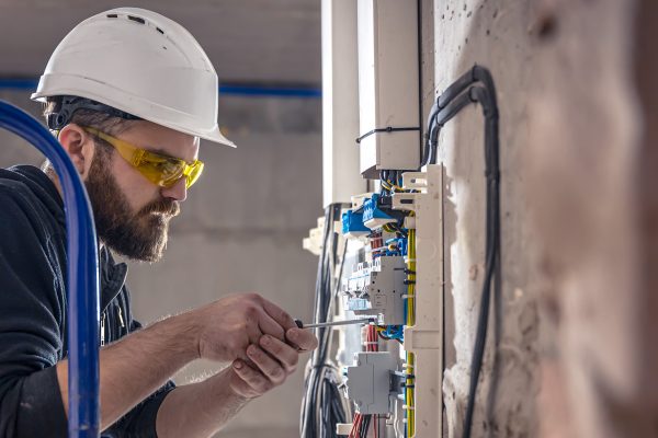 A male electrician works in a switchboard with an electrical connecting cable, connects the equipment with tools.
